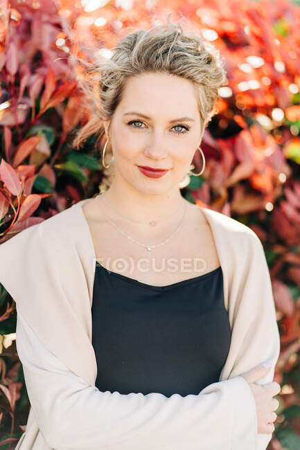 Happy blonde female in elegant dress and coat standing between trees and looking at camera — Stock Photo