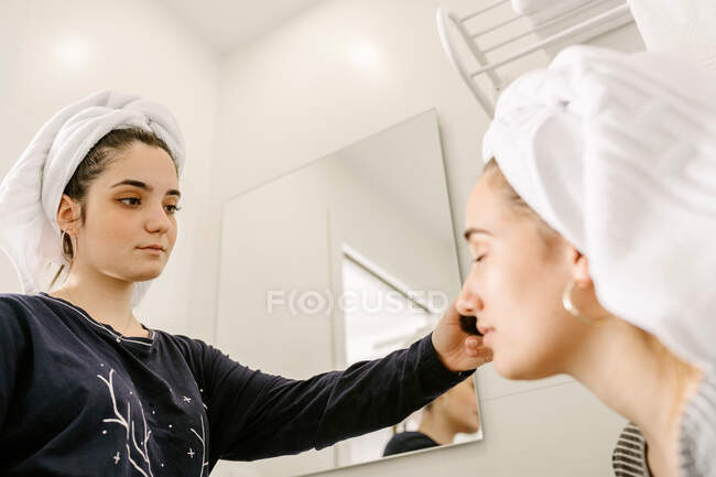 Снизу молодая этническая женщина в повседневной одежде и полотенце на голове наносит макияж на лицо лучшего друга, сидящего в ванной комнате с закрытыми глазами — стоковое фото
