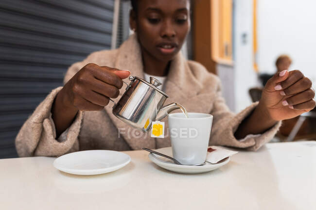 Обрежьте молодую афроамериканку в теплом пальто, наливая горячую воду из чайника в чашку, сидя за столом на открытой террасе кафе — стоковое фото