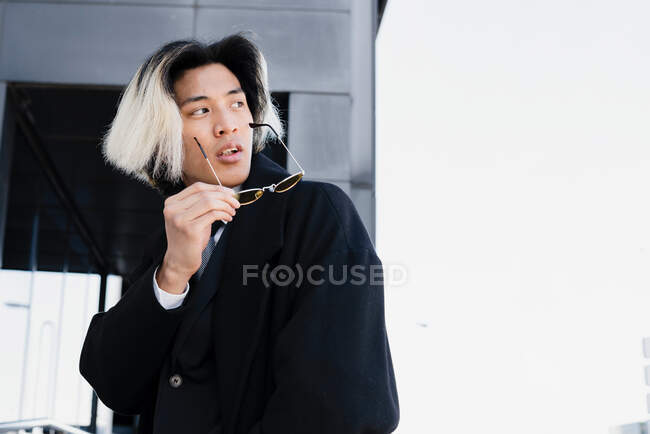 Giovane asiatico maschio esecutivo in tuta e occhiali da sole con le mani in tasca guardando altrove tra corrimano in città — Foto stock