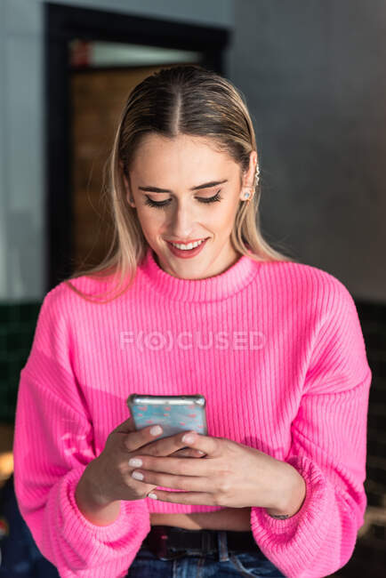 Содержание молодая женщина в розовом трикотажном свитере просматривает современный смартфон в светлом помещении — стоковое фото