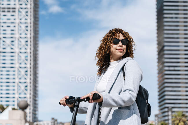 Optimistic young African American female with curly hair wearing blue coat and sunglasses standing with scooter on city street on sunny spring weather - foto de stock