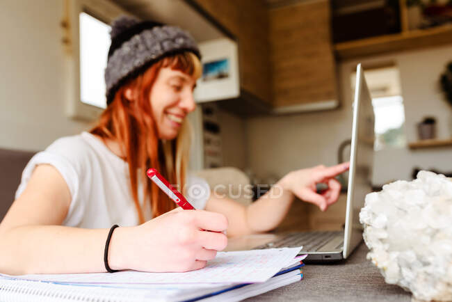 Side view of happy female traveler sitting at table in truck and taking notes while working on project remotely on laptop — Stock Photo