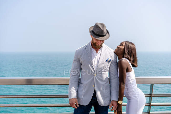 Happy young diverse couple wearing classy outfits standing together on city embankment against rippling blue sea on sunny day — Stock Photo