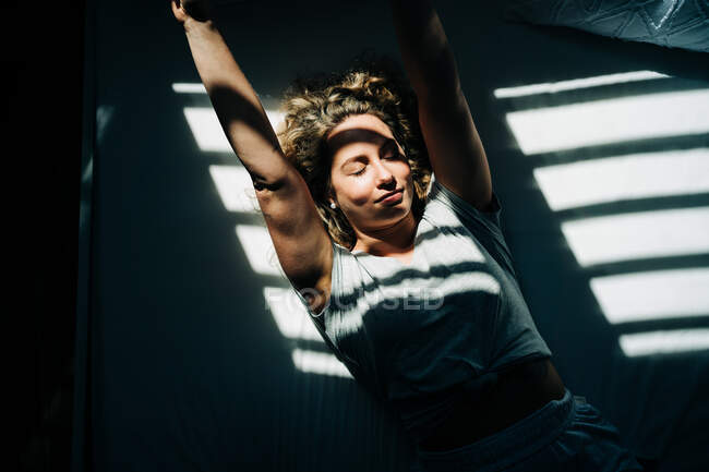 Зверху молода жінка з кучерявим світлим волоссям з закритими очима лежить на зручному ліжку в сонячний ранок — стокове фото