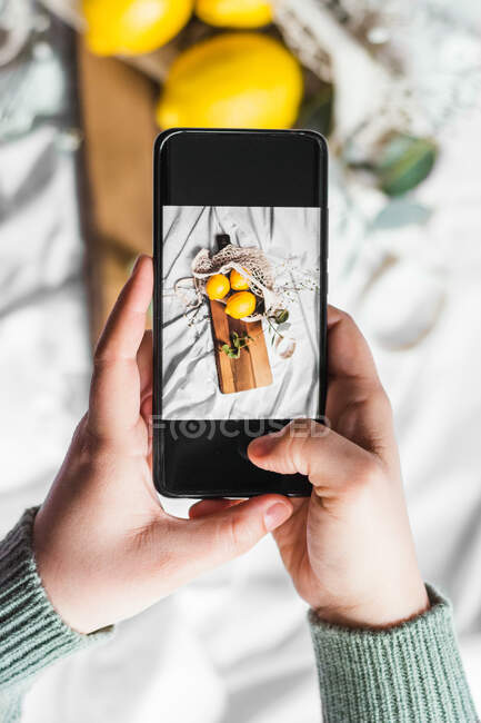 Top view of crop unrecognizable person touching screen of cellphone while taking photo of lemons on cutting board — Photo de stock