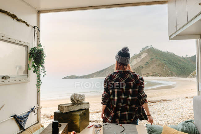 Rückansicht einer unkenntlich reisenden Kunsthandwerkerin, die neben einem LKW mit handgefertigten Accessoires vor dem Hintergrund von Meer und Bergen steht — Stockfoto
