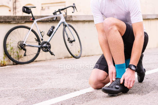 Crop unrecognizable male bicyclist in sports clothes and modern cycling shoes squatting on roadway against bike - foto de stock
