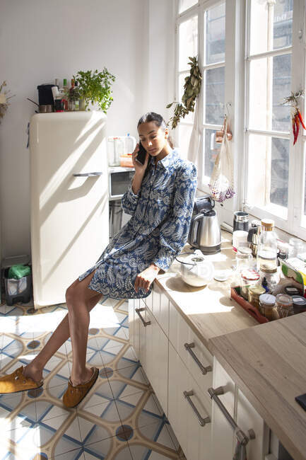 Полное тело стройная этническая женщина в повседневной одежде, разговаривающая по мобильному телефону и сидящая на современной кухне — стоковое фото