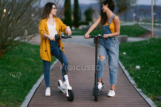 Cultivo alegre mejores amigas en ropa casual con scooters eléctricos mirándose unos a otros en la pasarela en la ciudad - foto de stock