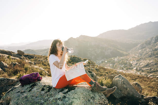 Side view of tired young female hiker with curly dark hair in casual outfit and backpack reading map and drinking water while resting on stone during trekking in mountainous valley — Stock Photo