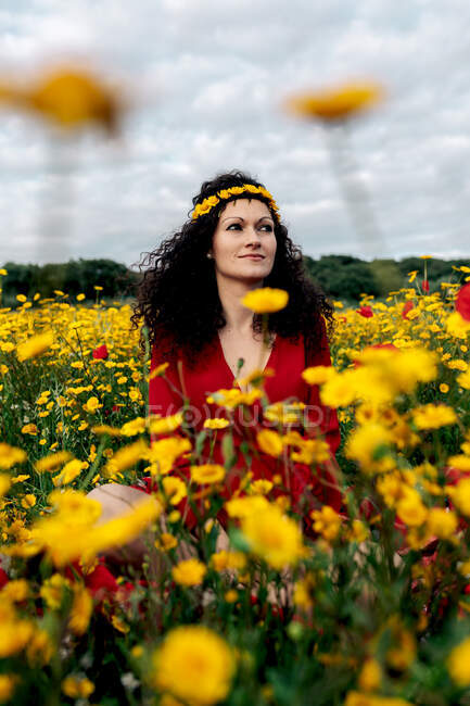 Зверху в захваті жінка в червоному сараї і з квітковою короною стоїть озираючись на квітуче поле з жовтими і червоними квітами, насолоджуючись теплим літнім днем — стокове фото