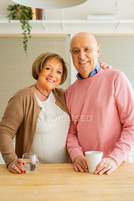 Zufriedenes Paar mittleren Alters steht mit Tassen voller Getränke in der Küche, während es sich umarmt und in die Kamera blickt — Stockfoto