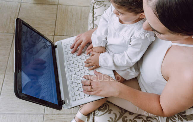 From above of ethnic woman with little girl on knees sitting in sofa at home and watching cartoon on netbook together — Stock Photo