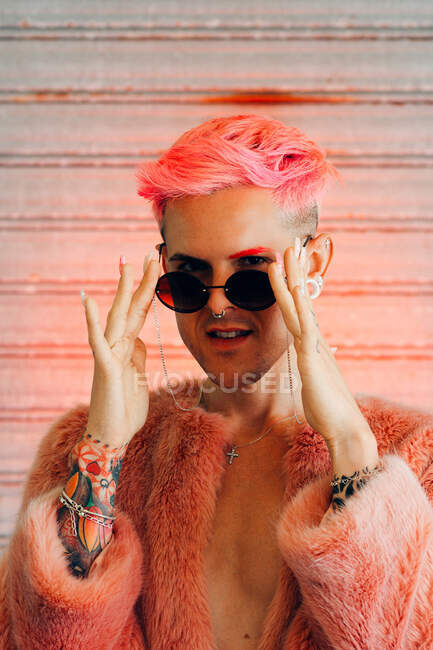 Молодий гей у стильному одязі з татуюванням, рожевим волоссям та манікюром з пухнастою курткою тримає сонцезахисні окуляри, дивлячись на камеру — стокове фото