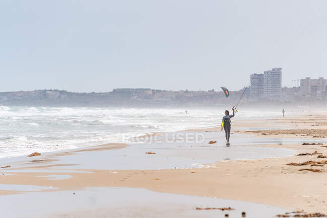 Спортсменка в гидрокостюме с надувным змеем прогуливается по песчаному берегу, глядя в сторону бурного океана — стоковое фото