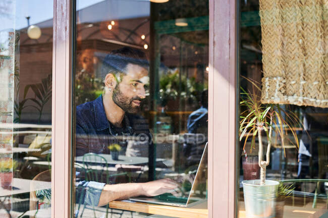 Através da visão de vidro do adulto étnico masculino remoto empregado trabalhando no netbook na mesa — Fotografia de Stock
