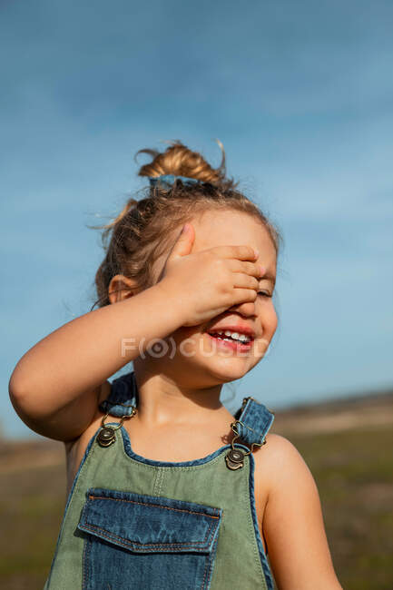 Delighted adorable little girl in overalls standing with hand on face in meadow and closed eyes — Stock Photo