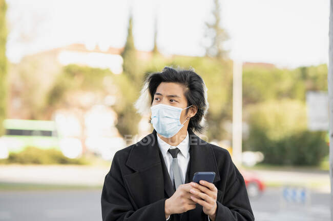 Молодой этнический мужчина исполнительный в одноразовой маске с сотовым телефоном глядя в город на размытом фоне — стоковое фото