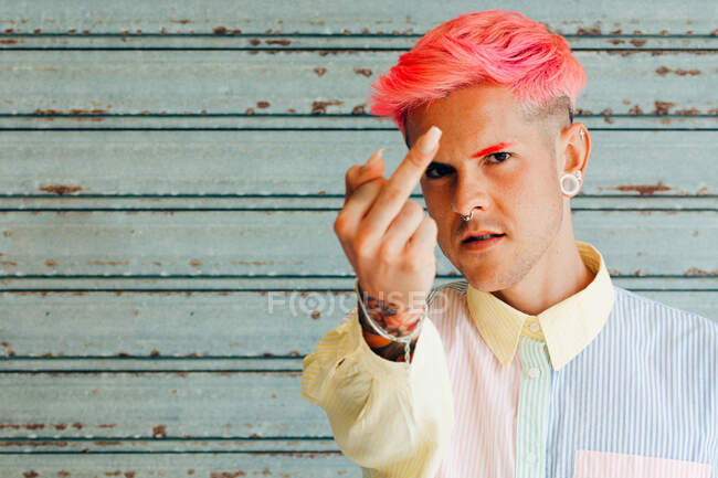 Гомосексуалист в полосатой рубашке с татуировкой и розовыми волосами демонстрирует средний палец, глядя в камеру — стоковое фото