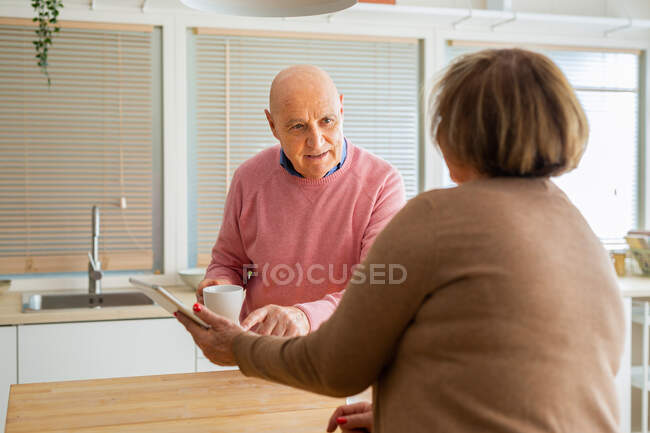 Мужчина средних лет разговаривает с женщиной с помощью планшета, стоя вместе на кухне дома — стоковое фото