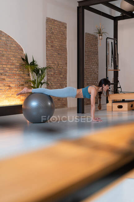 Vista lateral da fêmea em sportswear fazendo exercício na bola de ajuste durante o treinamento no ginásio pilates — Fotografia de Stock