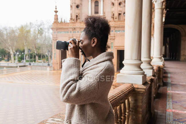 Vista lateral joven mujer afroamericana fotógrafa en abrigo cálido tomando fotos de edificios históricos de la ciudad en la cámara fotográfica moderna en el primer día de primavera - foto de stock