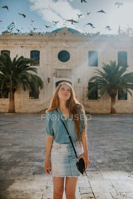 Conteúdo jovem viajante feminina em roupas casuais em pé contra o edifício de pedra medieval perto de pássaros voadores no dia ensolarado em Heraklion Creta — Fotografia de Stock