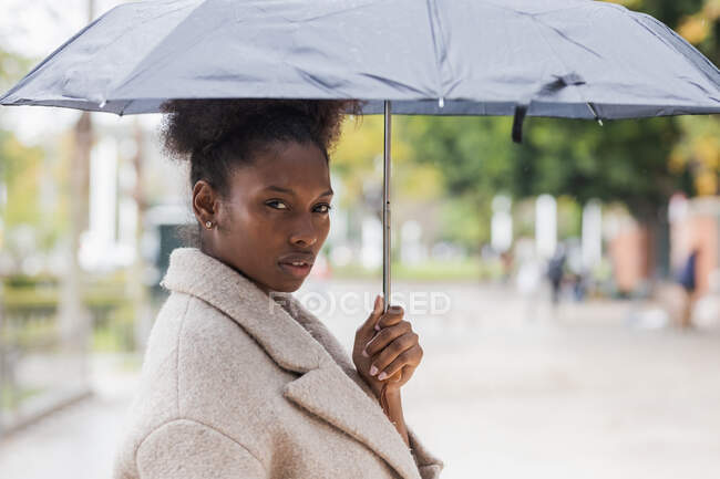 Молода африканська жінка в теплому пальто стоїть з парасолькою на сучасній вулиці міста і дивиться на камеру — стокове фото