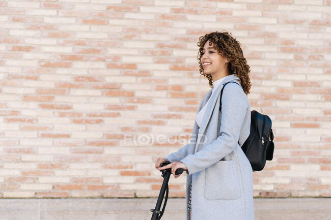 Content African American young female in casual wear riding scooter on paved sidewalk and looking away with smile — Fotografia de Stock
