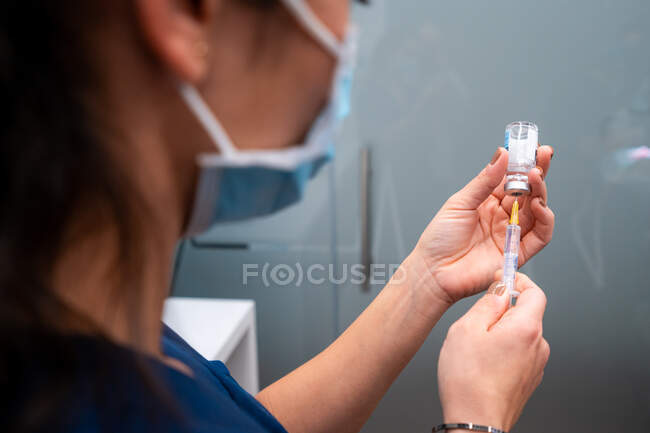 Вид сбоку обрезанных неузнаваемых женщин-ветеринаров в тканевой маске для лица, принимающих лекарство с помощью инъектора из бутылки — стоковое фото