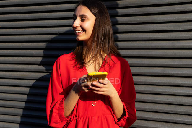 Young content female in red wear chatting on mobile phone in sunlight on gray background — Stock Photo
