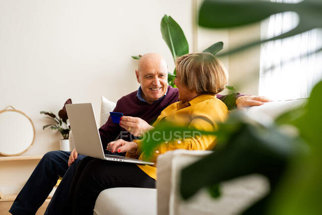 Casal maduro alegre fazendo pagamento com cartão de plástico durante compras on-line via laptop em casa — Fotografia de Stock
