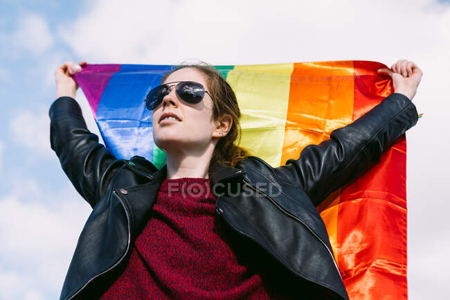 De baixo encantada lésbica fêmea de pé na rua com bandeira do arco-íris LGBT balançando no vento e olhando para longe contra o céu nublado — Fotografia de Stock