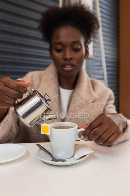 Crop young African American female in warm coat pouring hot water from teapot into cup while sitting at table on outdoor cafe terrace — Stock Photo