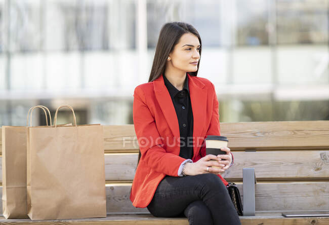Une cliente branchée assise sur un banc avec des sacs en papier et une boisson à emporter après avoir fait du shopping en ville et regardé ailleurs — Photo de stock