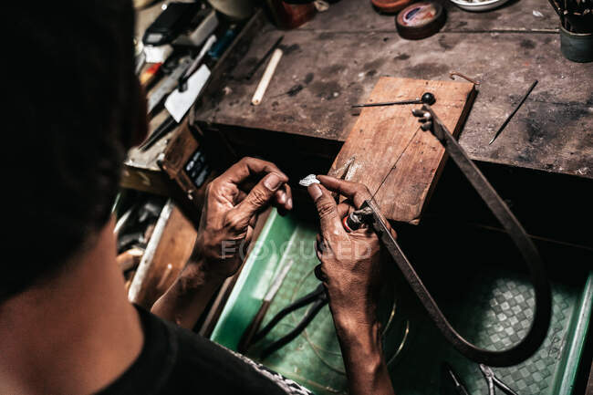 Von oben gesichtsloser Handwerker mit Metallpiercing-Säge und Silberstück bei der Arbeit am schäbigen Schreibtisch — Stockfoto