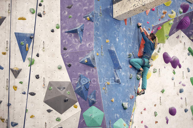Vista laterale di una donna forte nella parete di arrampicata con imbracatura di sicurezza durante l'allenamento in palestra di boulder — Foto stock