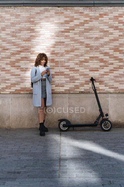 Happy young African American female with curly hair wearing blue coat browsing on mobile phone while standing against brick wall and looking at screen with smile — Stock Photo