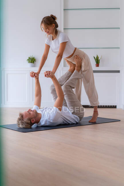Side view of boyfriend lifting girlfriend while doing acro yoga together at home and holding hands — Stock Photo