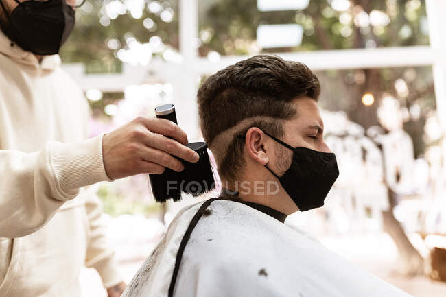 Male hairdresser in cloth face mask with brush removing hair from cape of client in barbershop — Foto stock