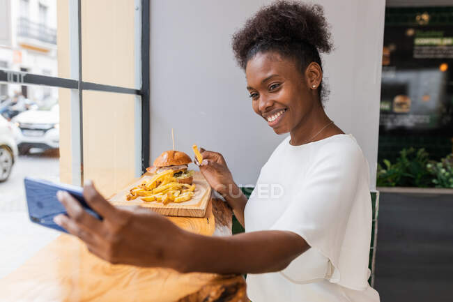Vista lateral alegre jovem afro-americana em blusa branca desfrutando de deliciosas batatas fritas e hambúrguer enquanto toma selfie no smartphone no restaurante fast food — Fotografia de Stock