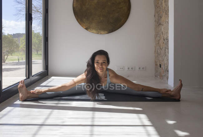 Flexível feminino realizando Upavista Konasana pose ao tocar pernas no tapete de ioga em casa olhando para a câmera — Fotografia de Stock