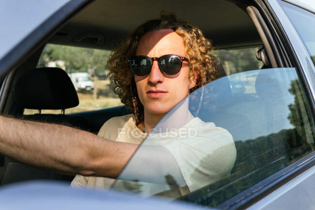 Ernster junger Mann mit stylischer Sonnenbrille blickt durch das geöffnete Autofenster in die Kamera, während er am Fahrersitz sitzt — Stockfoto