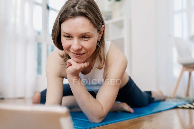 Deleitado delgado hembra acostado en la estera y tableta de navegación al elegir la lección en línea para practicar yoga - foto de stock
