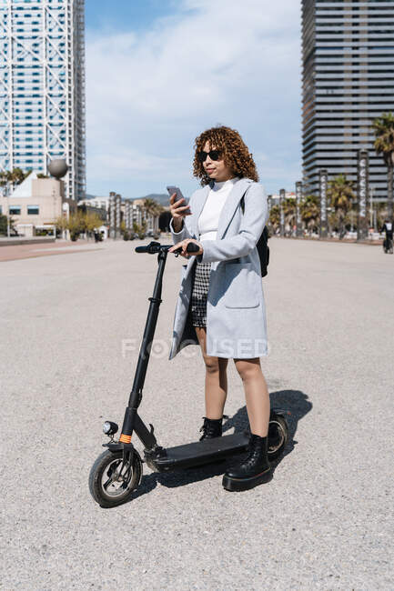 Full body young African American female in coat and sunglasses standing on city street with scooter and browsing mobile phone on sunny spring day — Foto stock