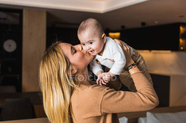 Seitenansicht der entzückten Mutter, die ihr entzückendes lächelndes Baby wirft und küsst, während sie zu Hause zusammen Spaß hat — Stockfoto