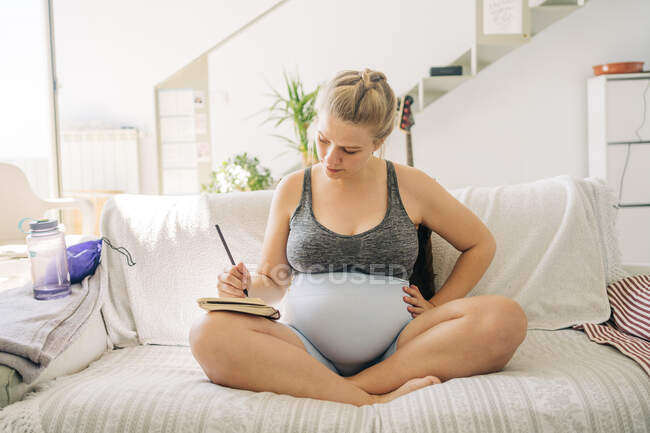 Молода вагітна жінка в спортивному одязі приймає нотатки в порядку денному, сидячи з схрещеними ногами на дивані у вітальні — стокове фото