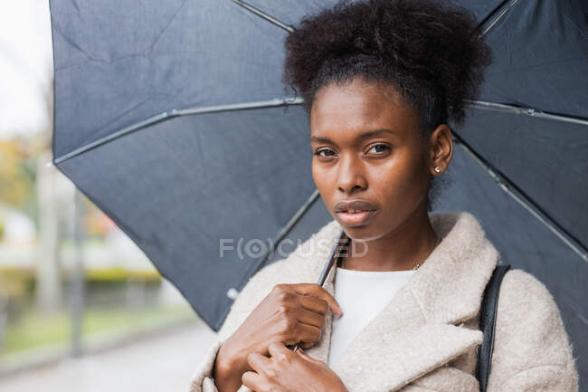 Joven mujer afroamericana de moda con abrigo cálido de pie con paraguas en la calle de la ciudad moderna y mirando a la cámara - foto de stock