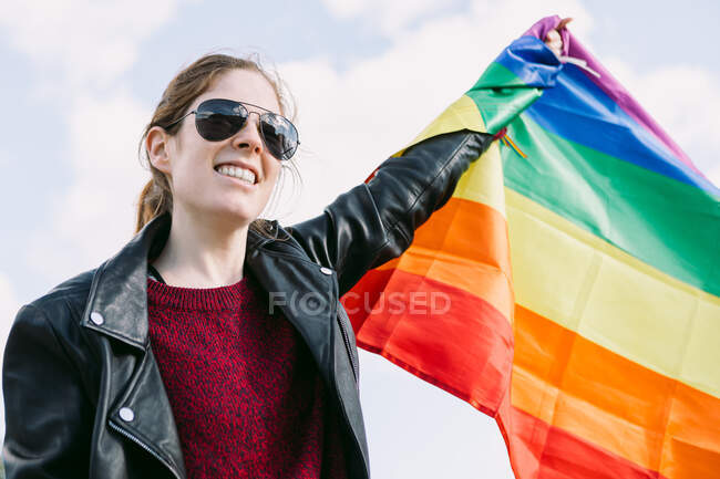 Снизу восторженная лесбиянка, стоящая на улице с радужным флагом ЛГБТ, развевающимся на ветру и смотрящим в сторону облачного неба — стоковое фото